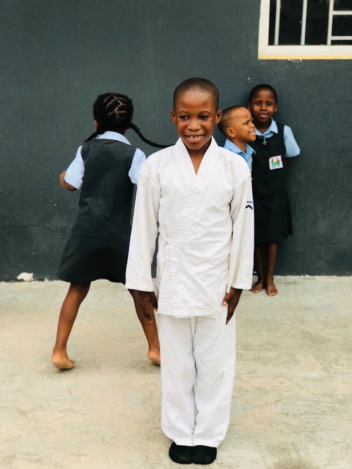 A smiling boy in a karate gi stands with friends in school uniforms against a dark wall.